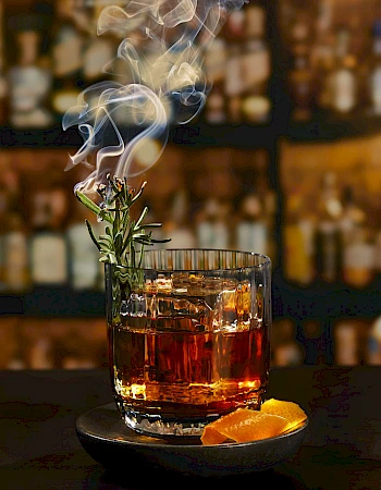 A smoky rosemary Old Fashioned with orange twist on a dark bar counter, glass steaming against a blurred liquor shelf.